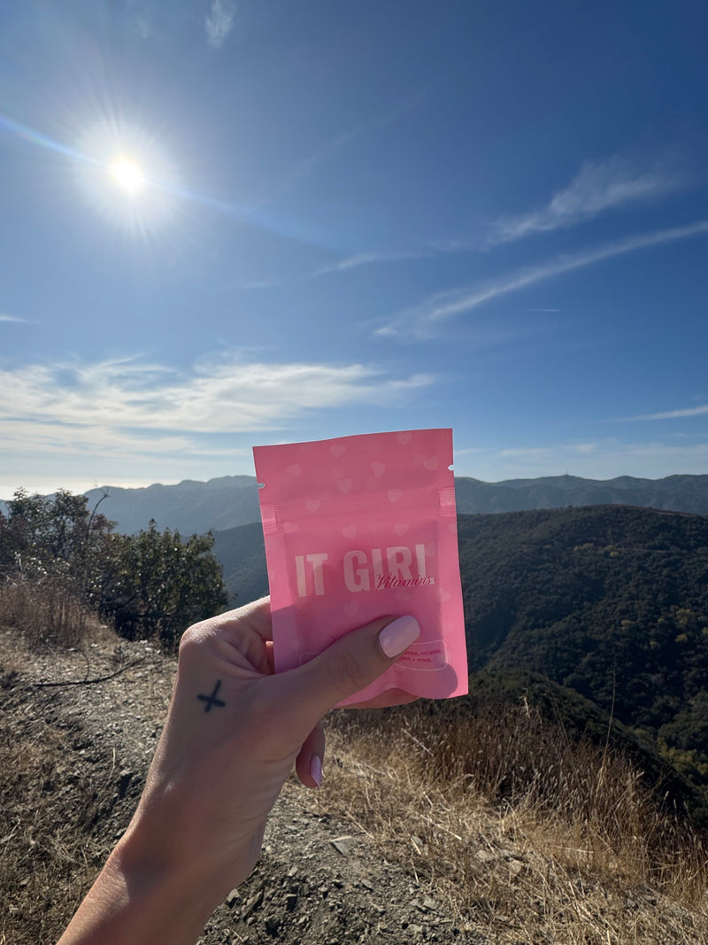 Hand holding a pink It Girl pouch on a sunny mountain trail.