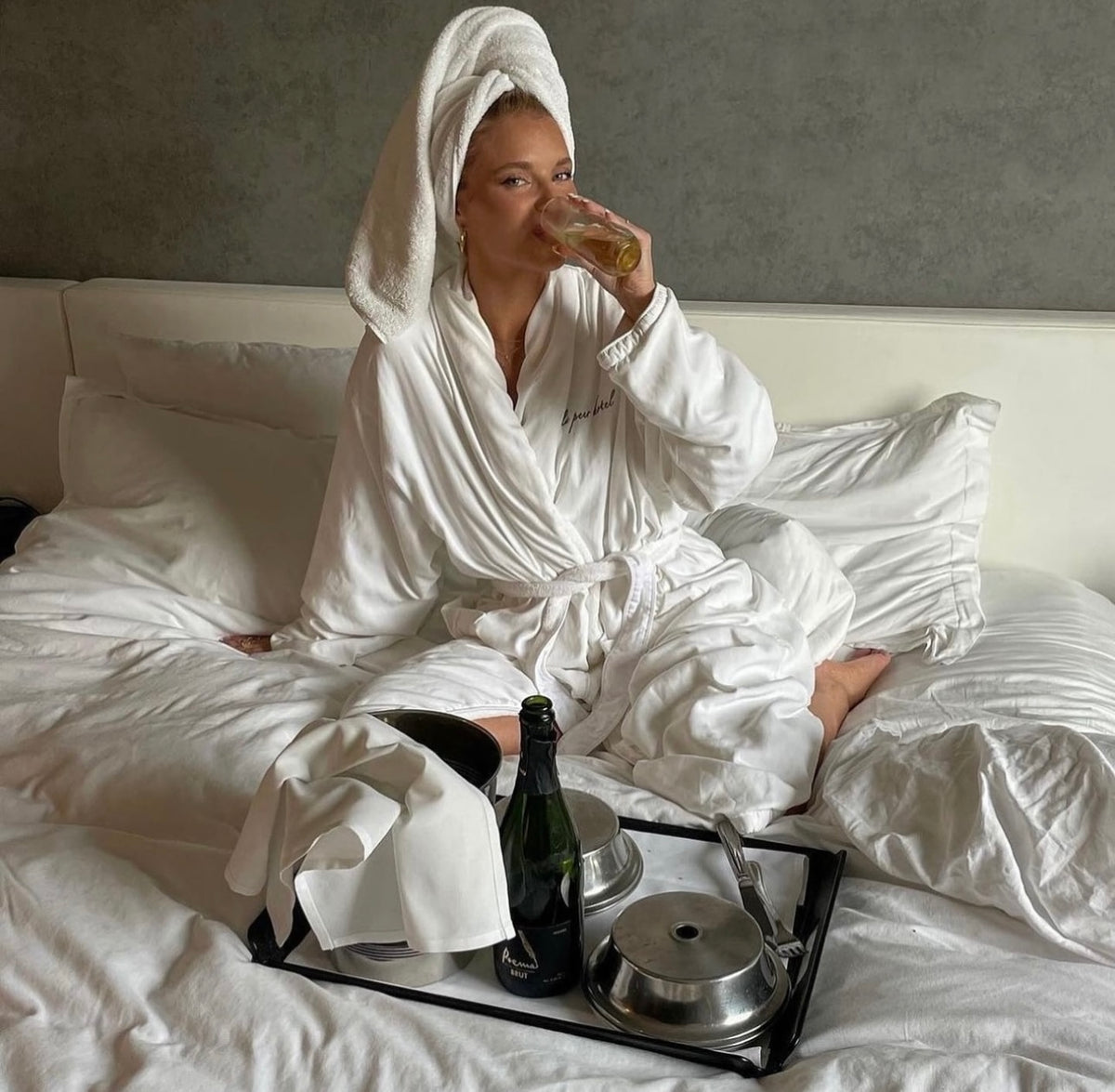 Woman in a white robe and towel drinking from a glass while sitting on a bed.