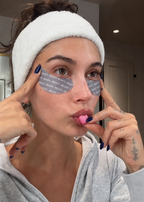 A woman wearing a white textured headband and a gray zip-up hoodie applies eye patches with her fingers while holding a small pink heart-shaped item to her lips. She is in a real indoor room with a door and ceiling light visible in the background.