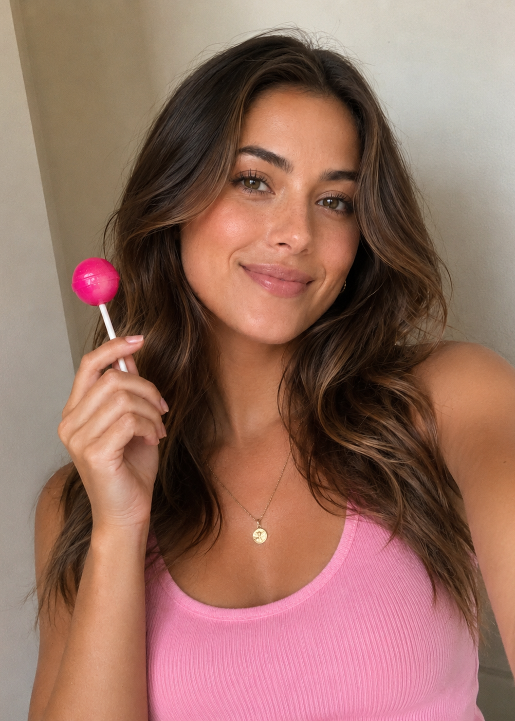 A woman with long wavy brown hair holds a bright pink lollipop while wearing a pink ribbed tank top and a small gold pendant necklace, photographed in a close-up indoor setting.