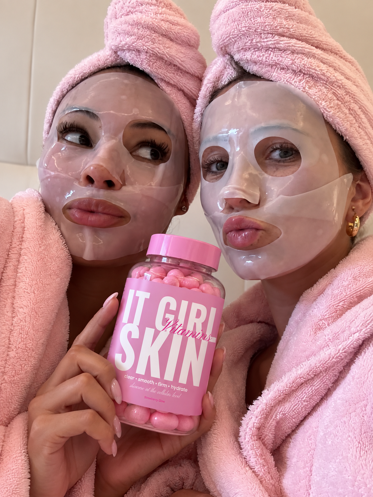 Two women wearing face masks and pink towels with an It Girl Skin bottle.