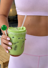 Woman in lavender activewear holding a green lollipop and iced green drink.