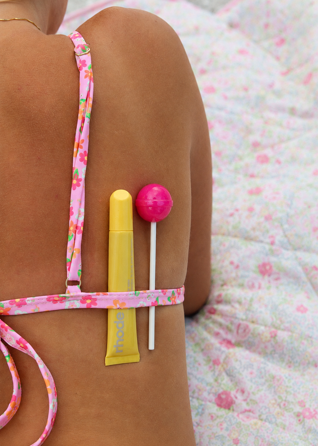 A close-up lifestyle shot of a person's bare back wearing a pink floral bikini top, with a yellow tube and a pink lollipop tucked into the straps, set against a matching pink floral blanket.